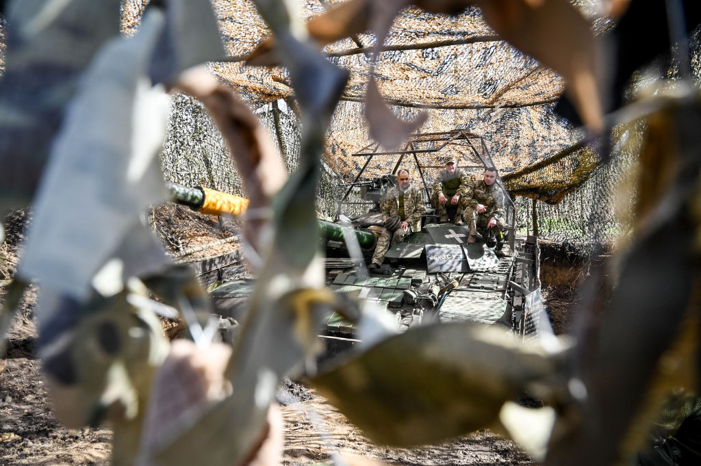 Tres militares sentados en un tanque de la 1ª Brigada de las Fuerzas Terrestres Ucranianas, visto a través de una red de camuflaje,  el 31 de marzo de 2024