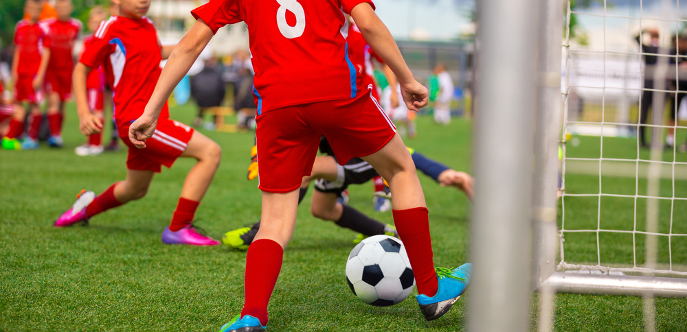 Young,Boy,Soccer,Player,Scoring,Goal,In,Match.,Junior,Level
