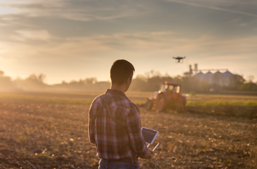 Attractive,Farmer,Navigating,Drone,Above,Farmland,With,Silos,And,Tractor