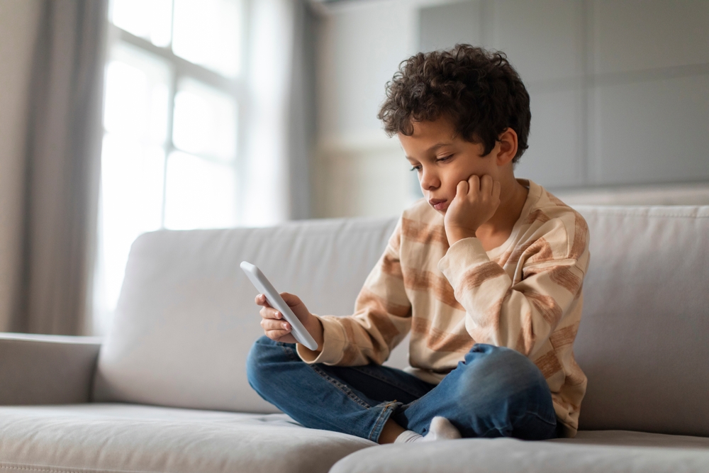 Bored,Black,Preteen,Boy,Sitting,On,Couch,And,Using,Smartphone