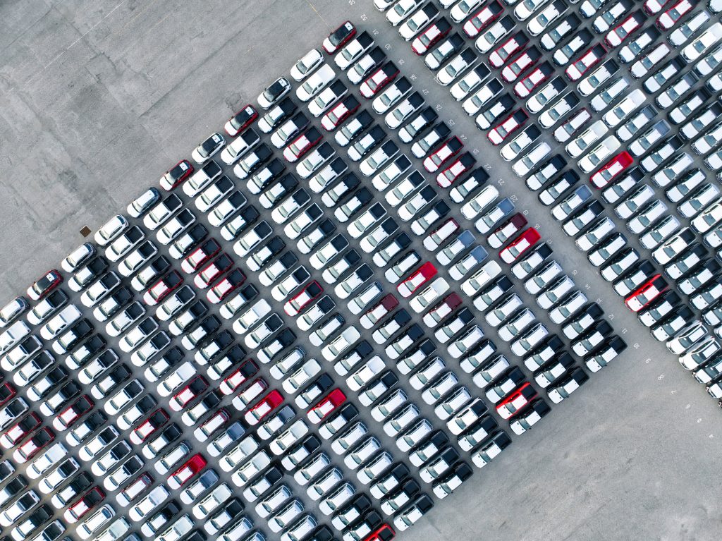 Aerial view of new cars stock at factory parking lot. Above view cars parked in a row. Automotive industry. Logistics business. Import or export new cars at warehouse. Big parking lot at port terminal